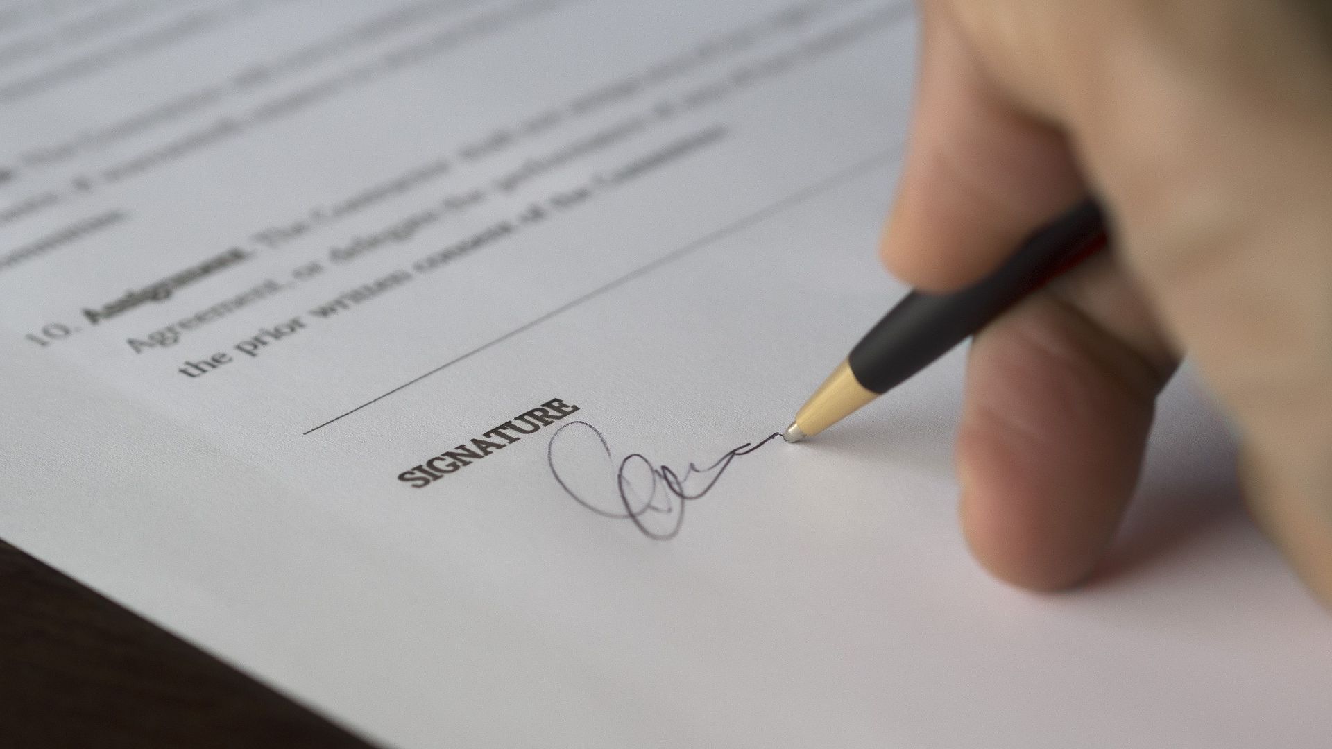 A hand signs a formal contract with a pen on a wooden desk.