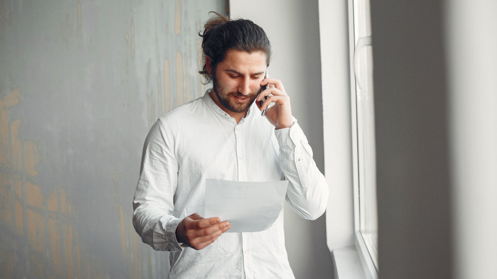 A young man in casual attire talks on the phone while holding a document indoors.