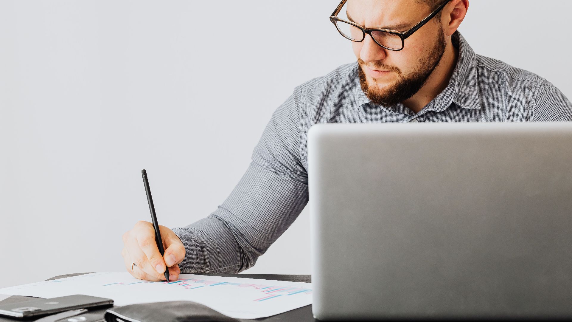 A focused man analyzing financial papers with a laptop in an office setting.