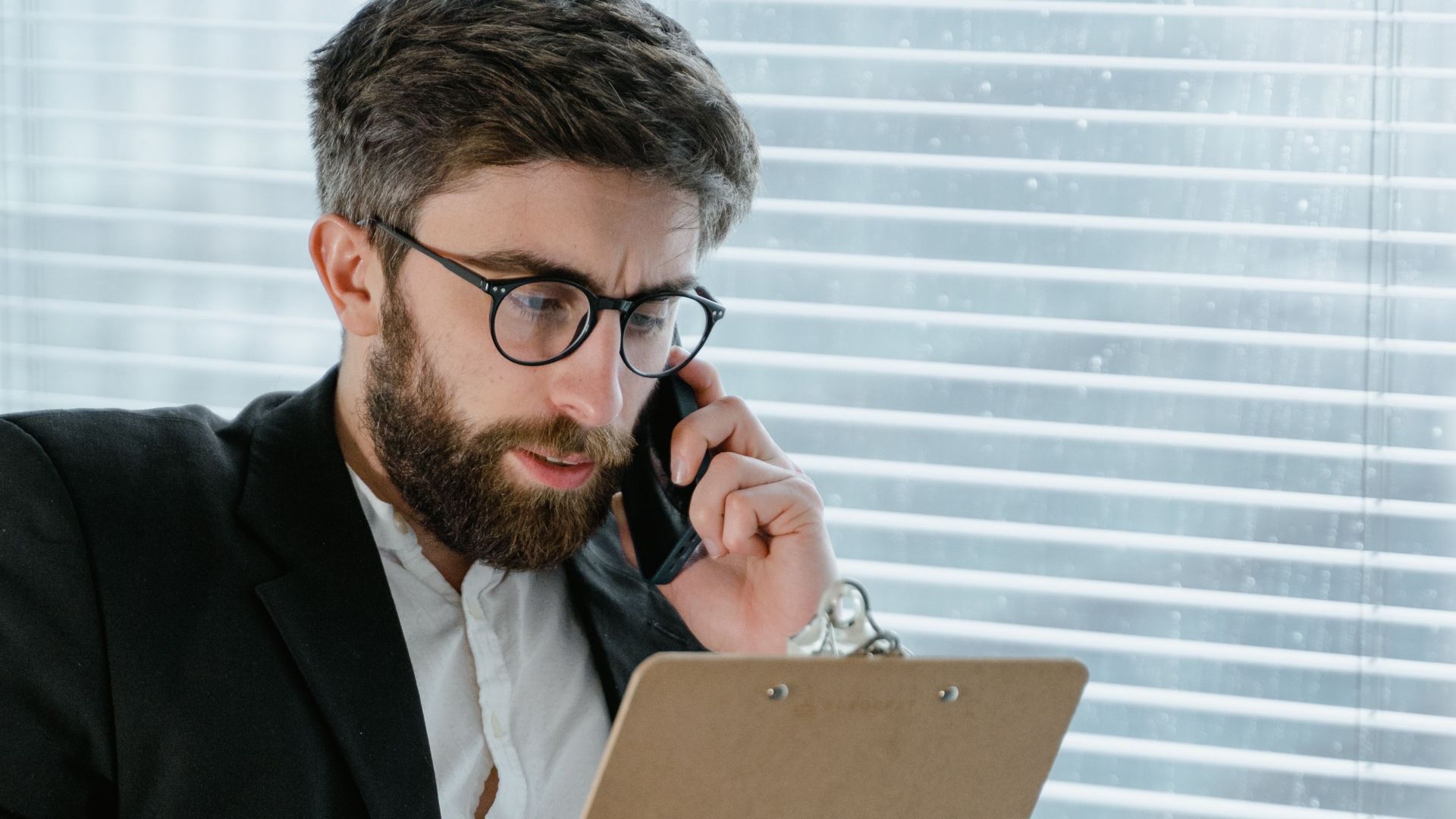 Professional businessman with a beard holding a clipboard while making a phone call indoors.