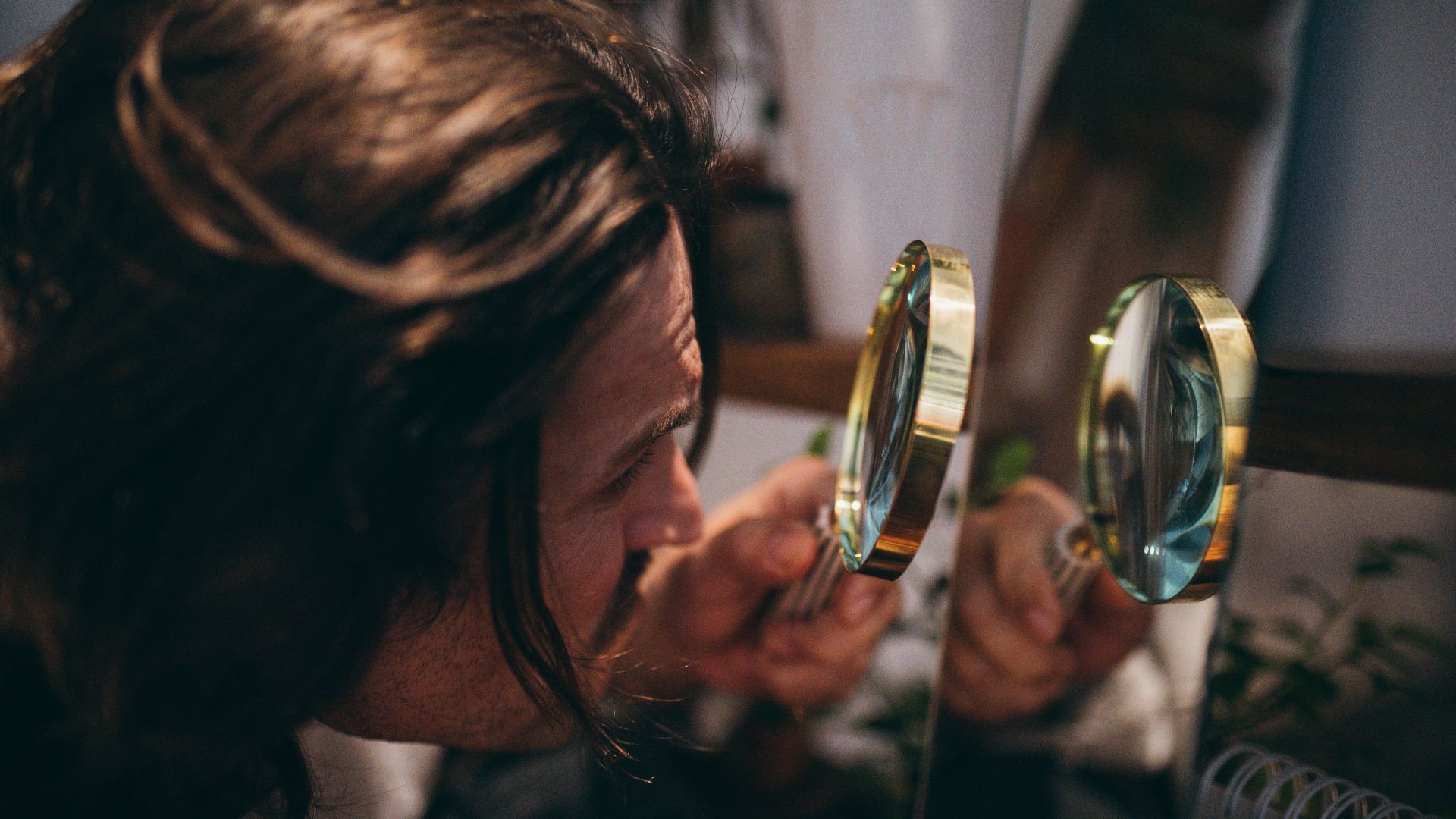 Close-up shot of a man with long hair using a magnifying glass to inspect his reflection in a mirror.