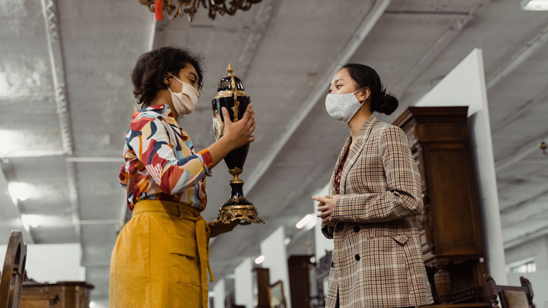 Two women in masks evaluating an antique vase in a shop, showcasing vintage fashion.