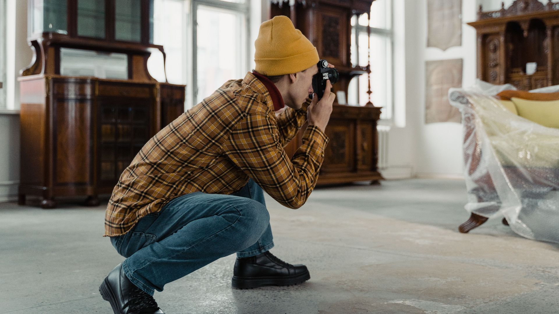 A person in a beanie photographs antique furniture inside a well-lit room.