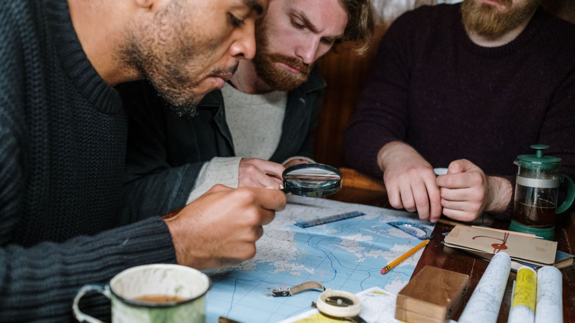 Three men are intensely discussing plans using maps and a magnifying glass, creating a scene of collaboration.