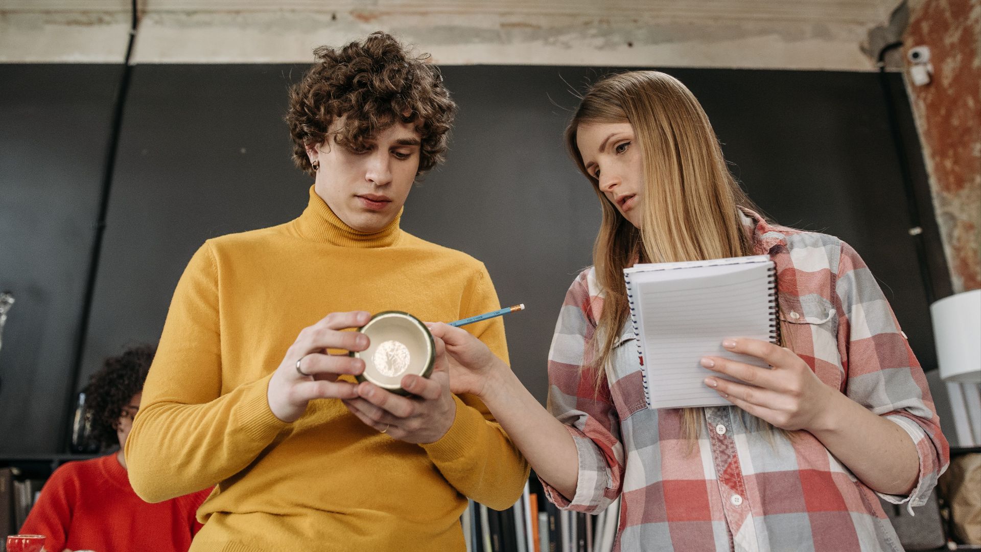 Young adults collaborating on ceramic design, featuring a man holding a cup and a woman with a notebook.