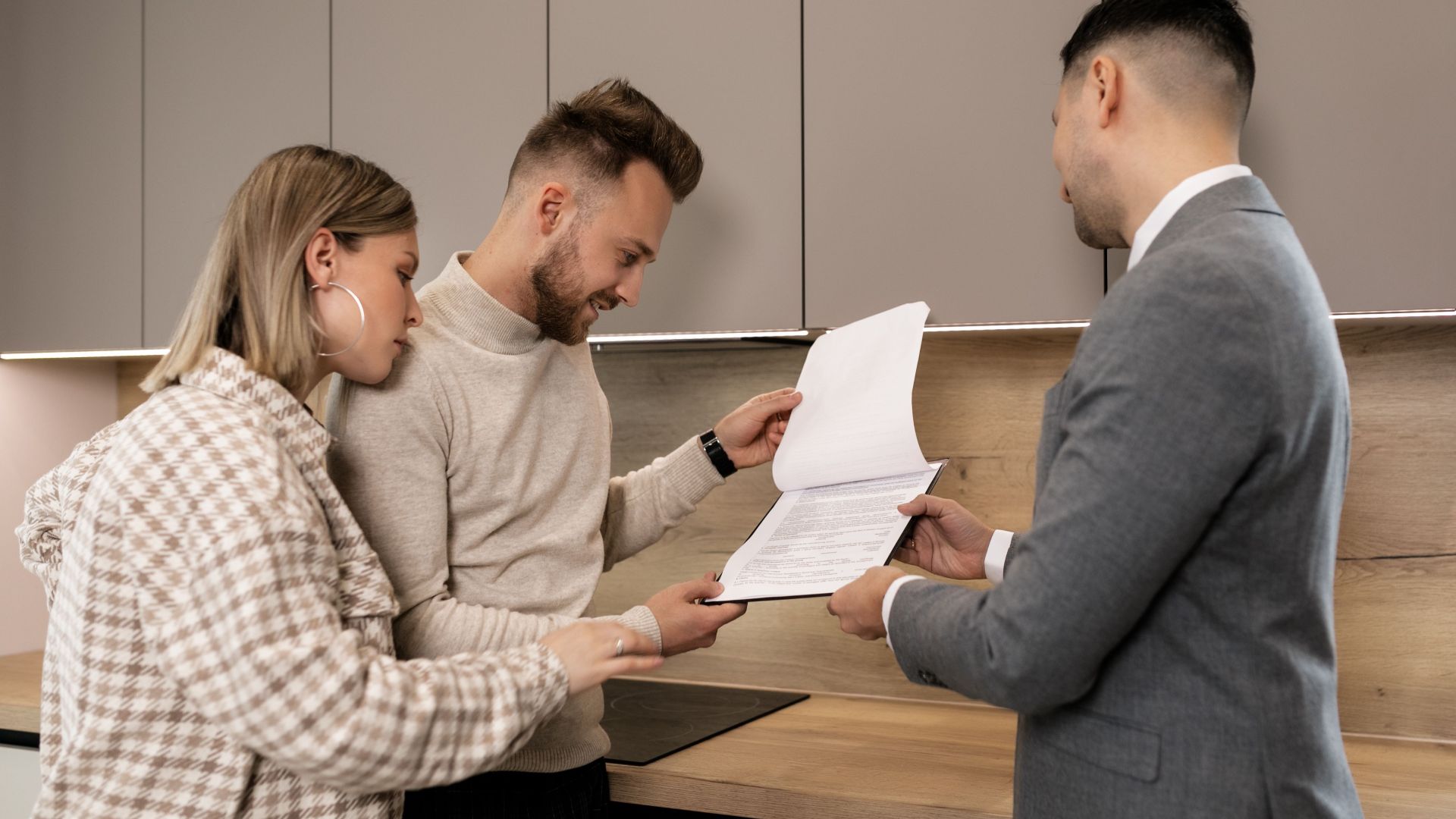 A couple reviews real estate documents with an agent in a modern indoor setting, discussing a potential property purchase.