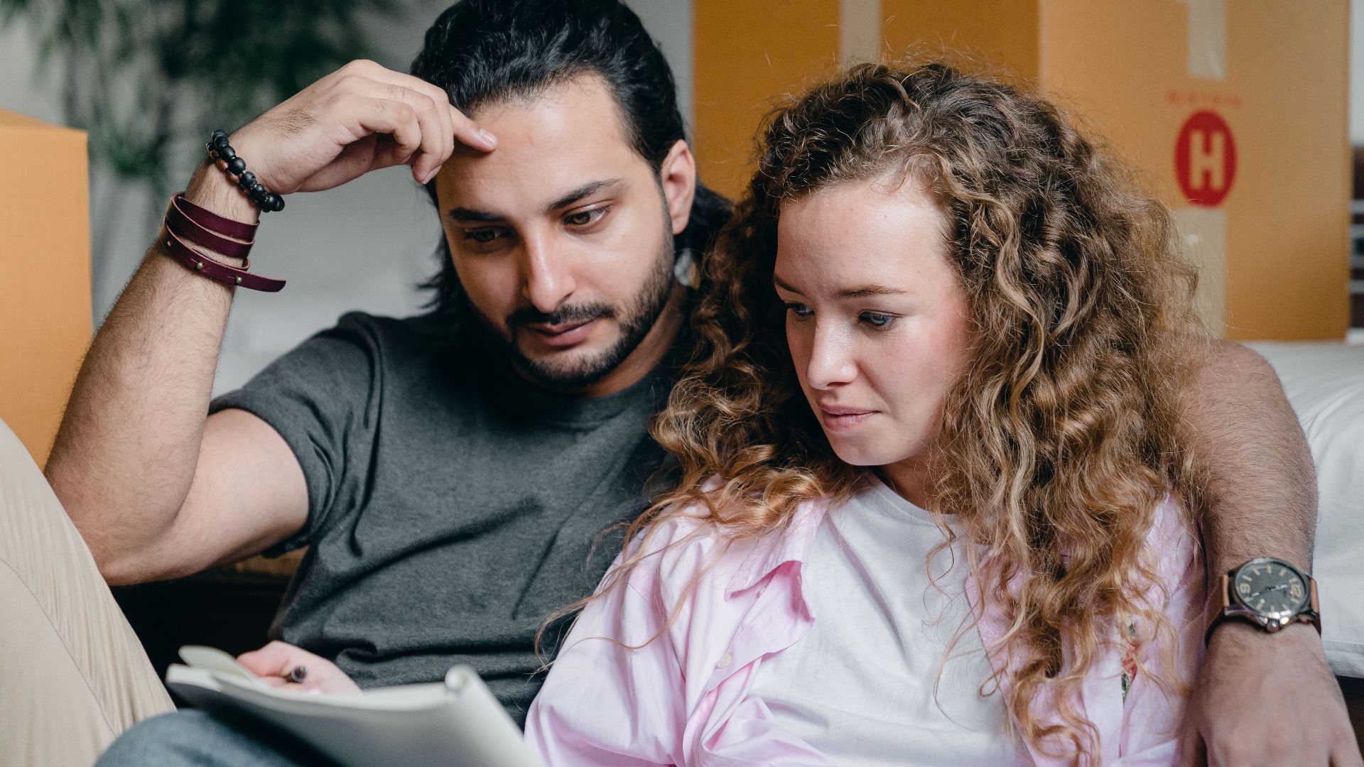 Focused male and female in casual wear sitting near boxes and looking through notebook thoughtfully while moving house