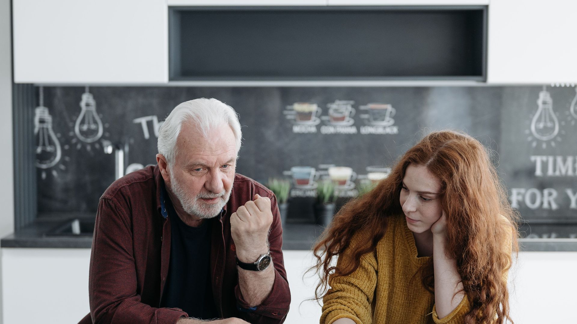 An elderly man and his granddaughter engage in a heartfelt conversation over coffee indoors.