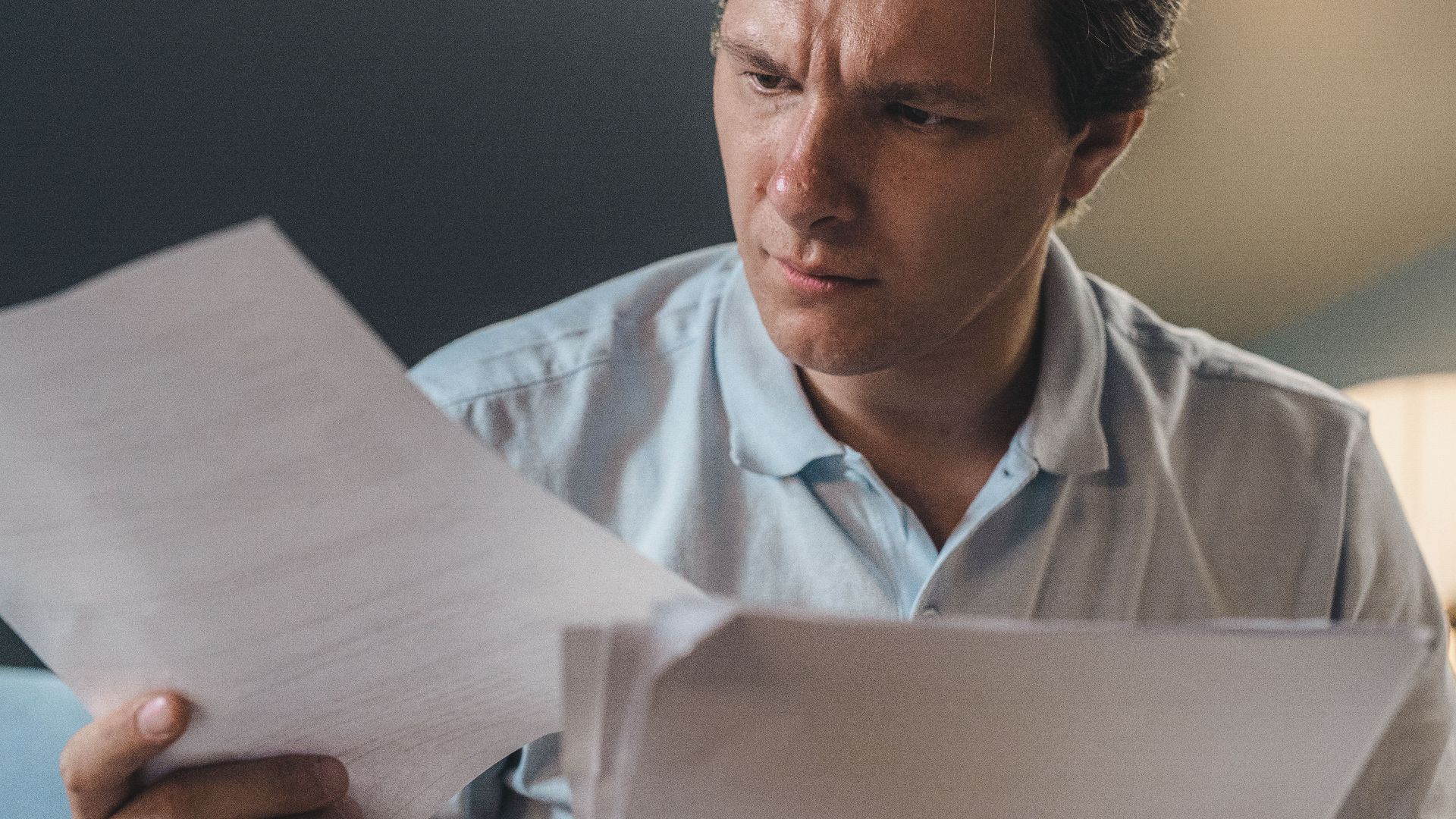 A man intently examines papers, seated indoors under warm lighting, focusing on his work.