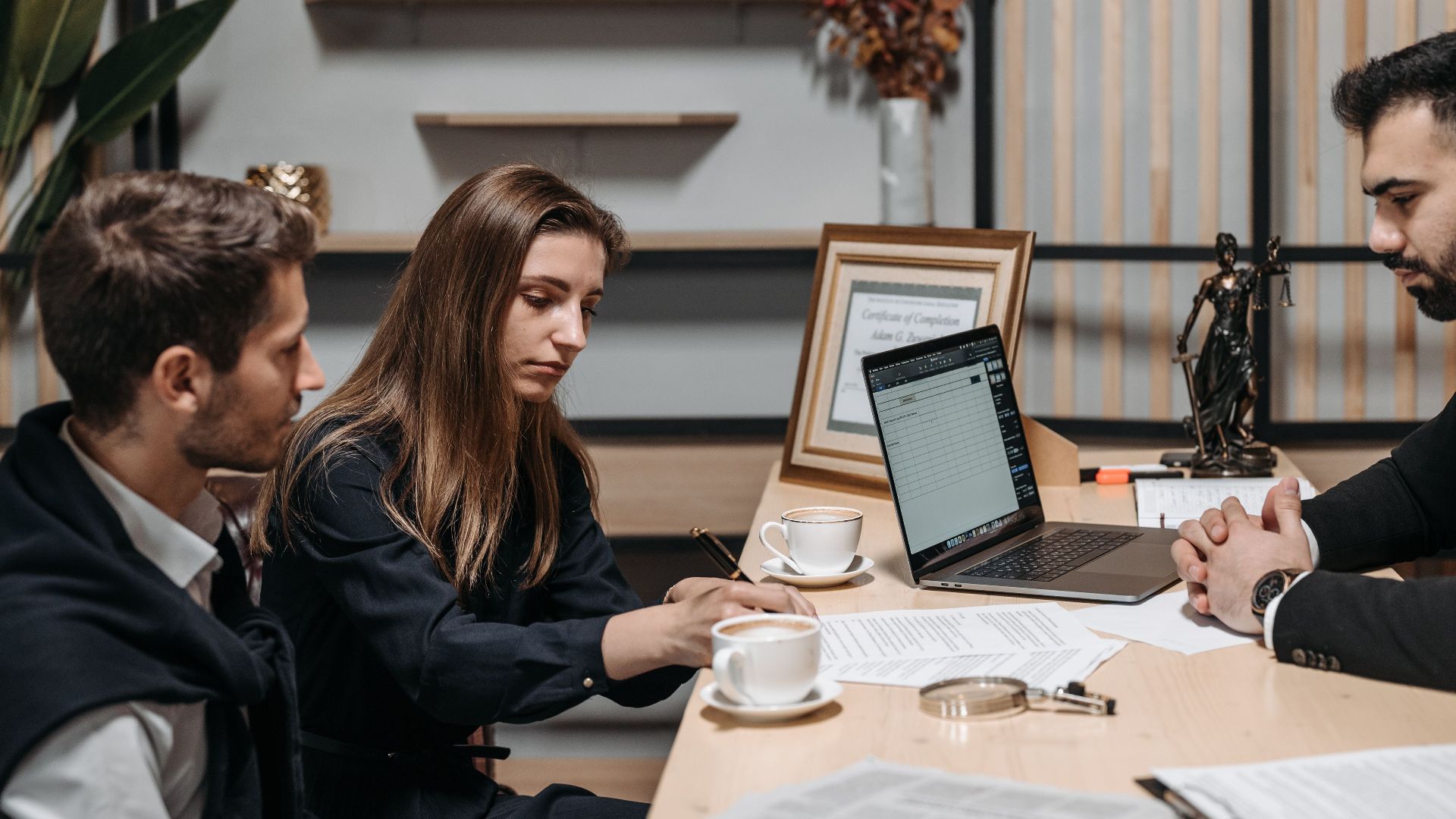 Three professionals discussing documents over coffee in a modern office setting.