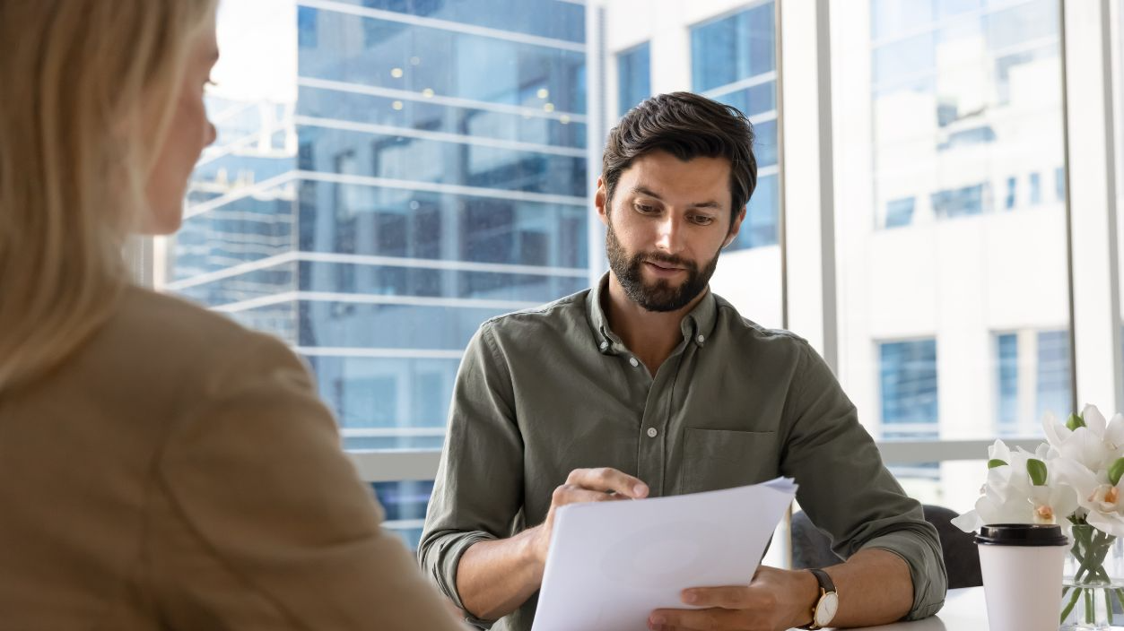 Shutterstock-2796542359, Professional business man attentively reads documents sits across table from woman in office, during formal business meeting or job interview, review data or resume paper. Human resources, staffing
