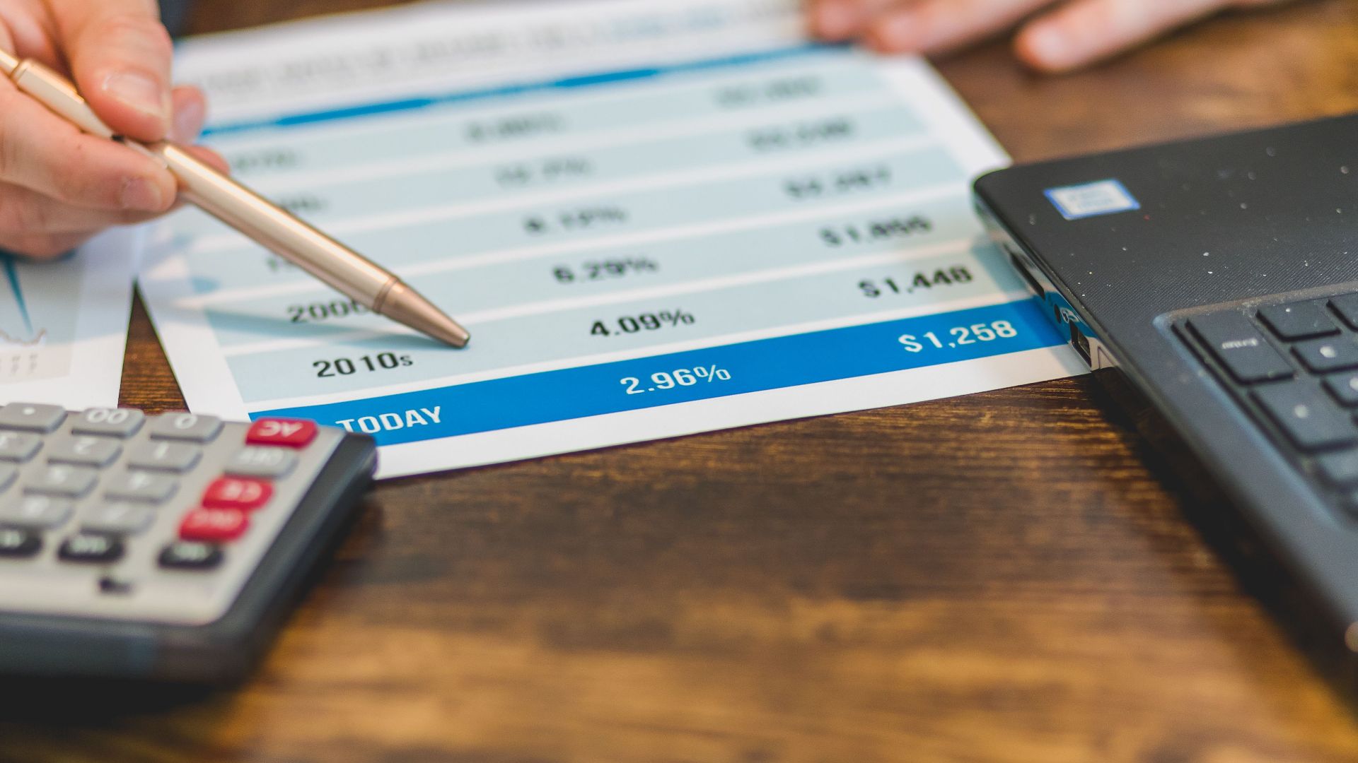 Close-up of a hand with pen analyzing financial rates on paper with a calculator and laptop nearby.