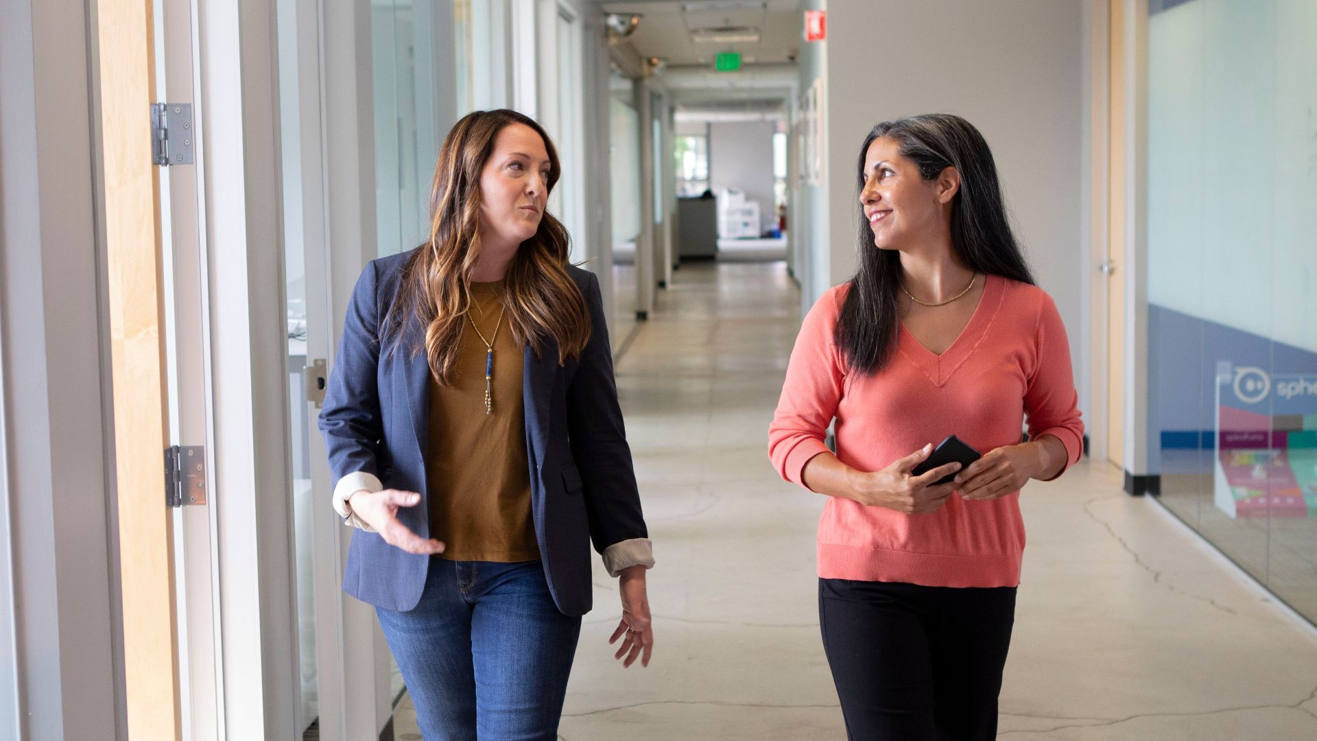Two professional women engaged in conversation while walking through a modern office corridor.