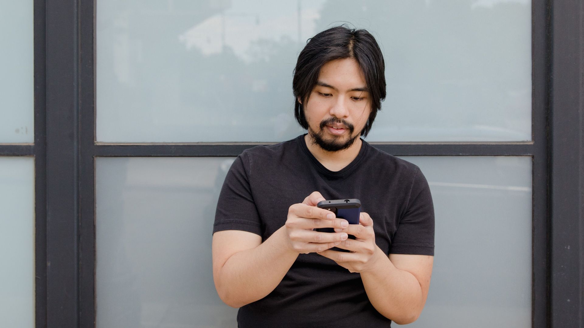 Asian man standing outdoors using phone against glass window backdrop.
