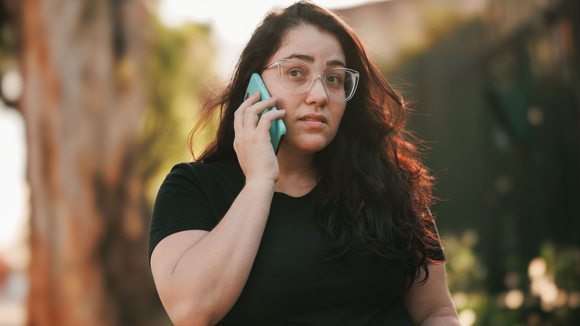 Woman with eyeglasses talks on smartphone in sunny park setting.