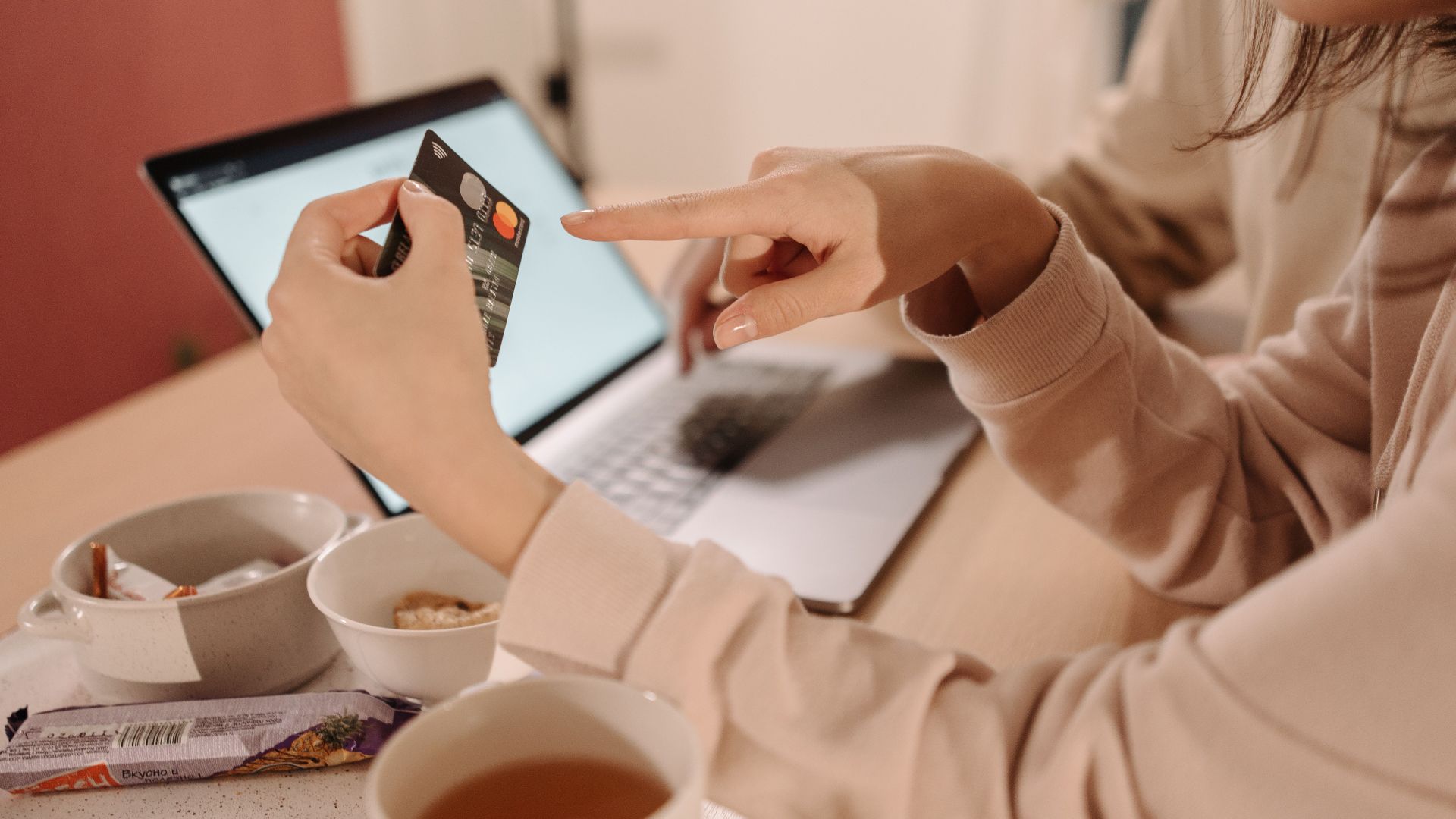 A woman using a credit card for online shopping while having tea and snacks on a table.