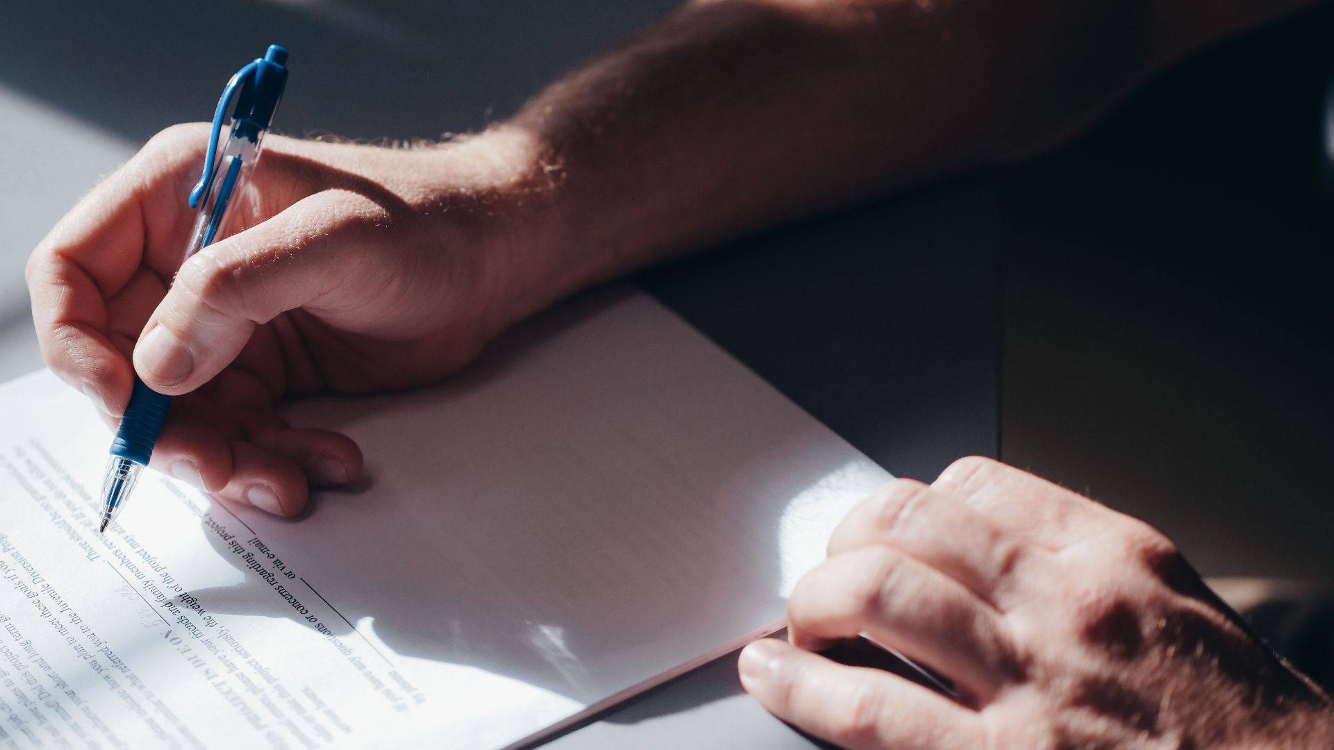 A close-up image of a person's hand signing a document with a blue pen in natural lighting.