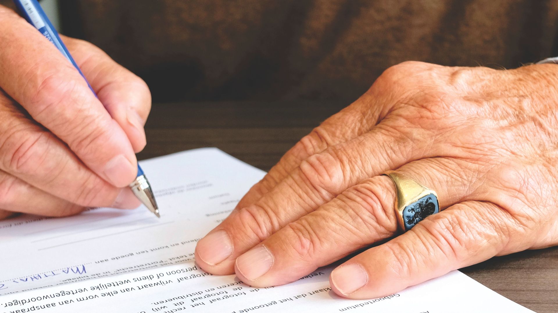 Close-up of a senior adult signing a legal document with a focus on hand and gold ring.
