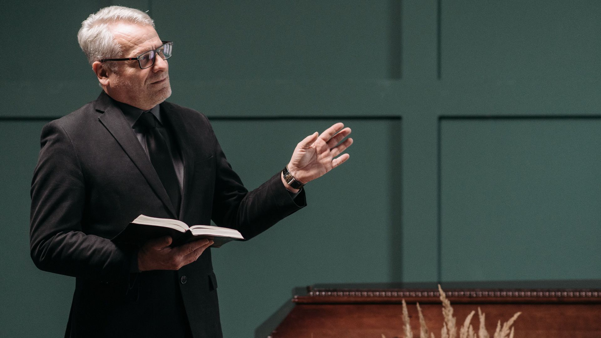 A pastor in black suit delivering a eulogy during a funeral ceremony indoors with a coffin.