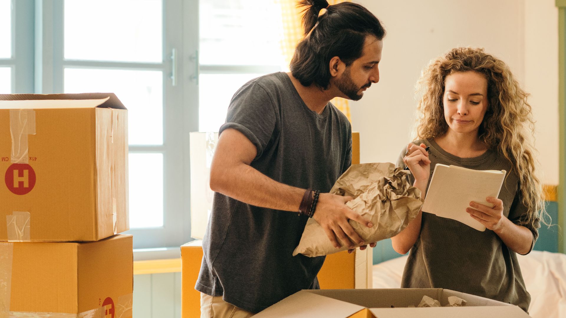 Cheerful young man and woman smiling while unpacking carton boxes with belongings in new apartment during relocation and looking at paper
