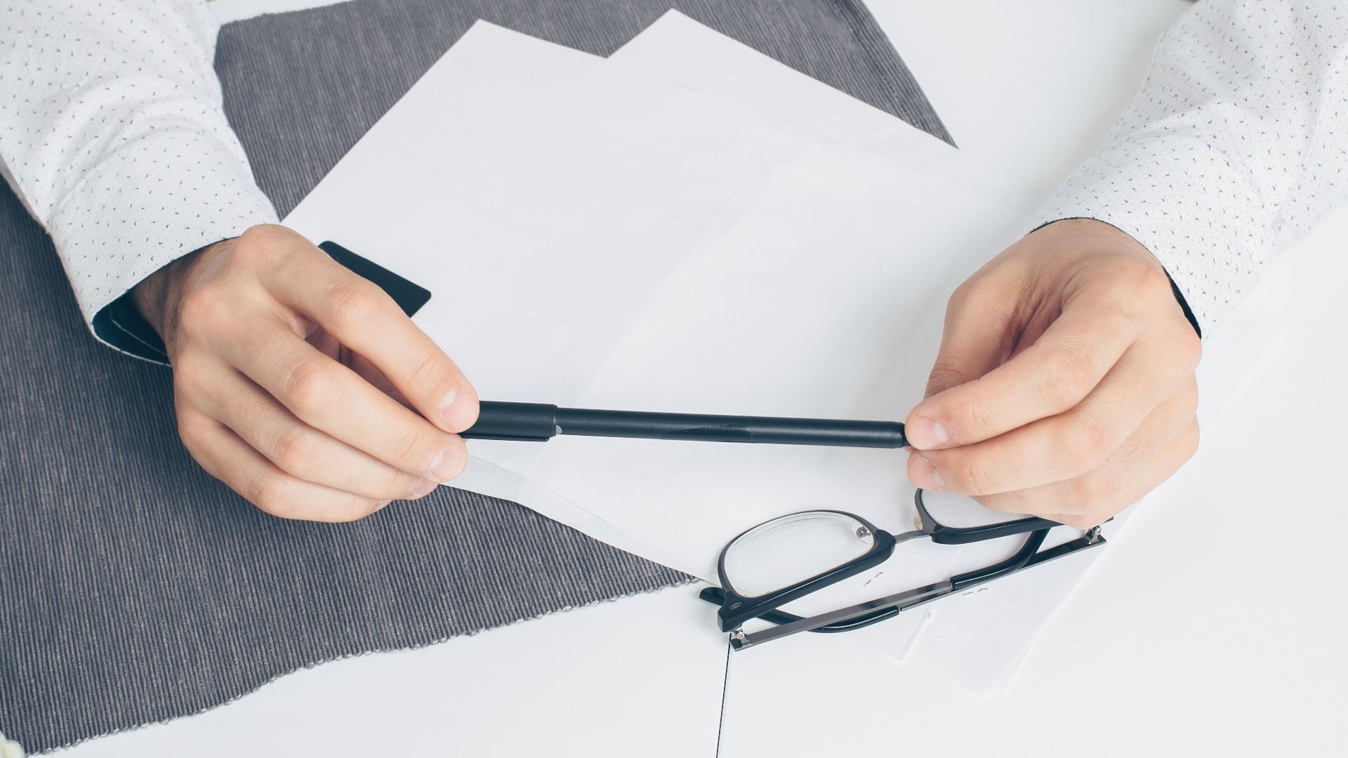 Close-up of hands holding a pen with papers and eyeglasses on a white table.