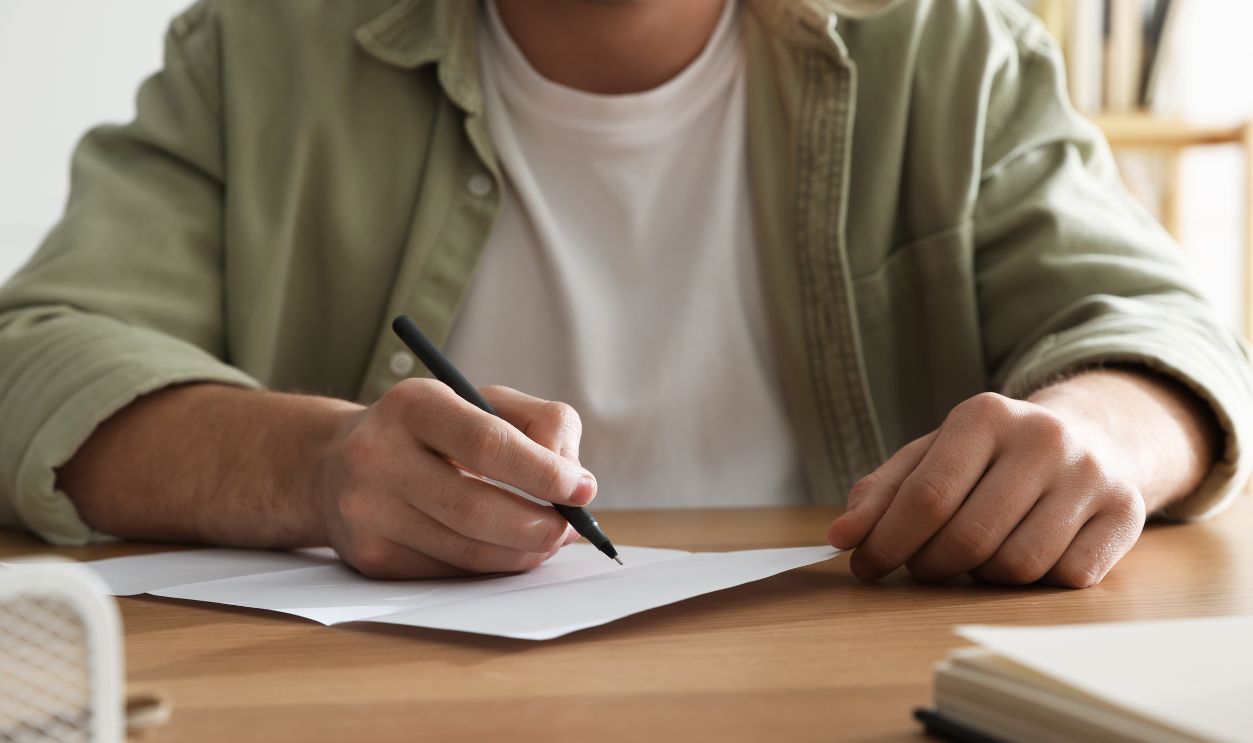Man writing letter at wooden table indoors, closeup