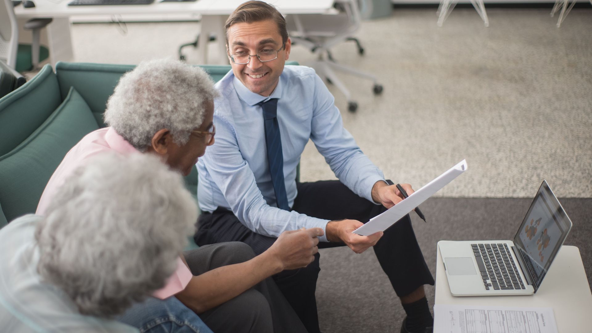 Consultant discussing financial plans with senior clients in a modern office setting, using documents and a laptop.