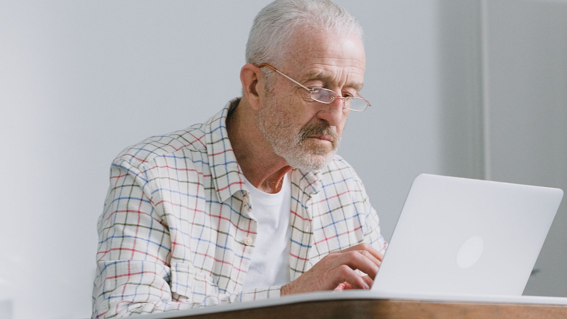 Elderly man focused on typing on laptop indoors. Natural lighting, modern ambiance.