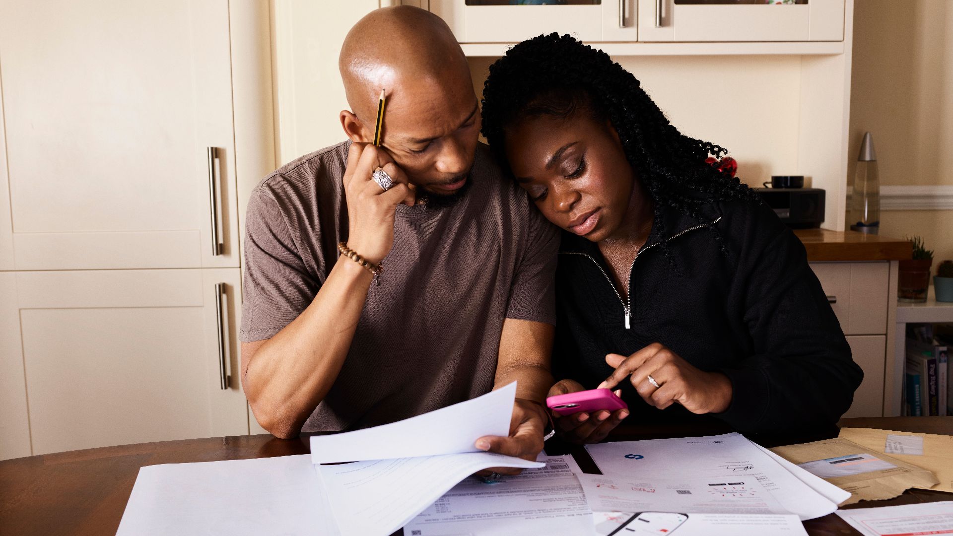 A couple sits at a table managing domestic finances, evaluating documents and using a smartphone.