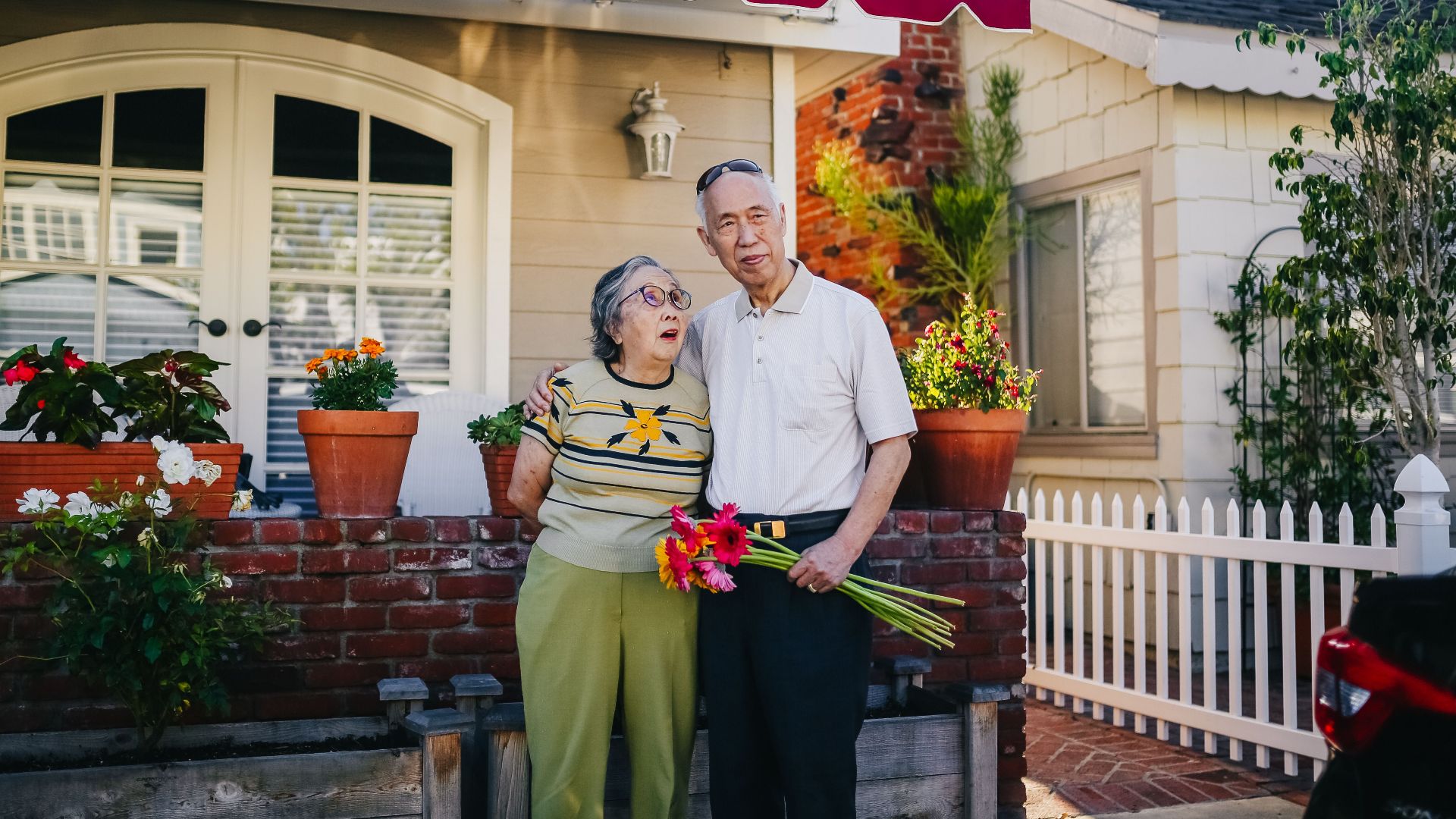 Elderly Asian couple embracing outside their cozy home with flowers, representing love and togetherness.