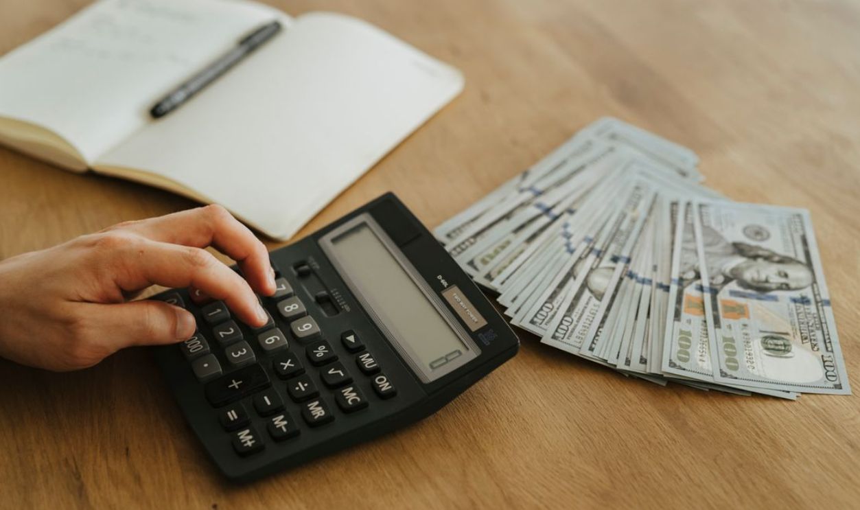 Close-up of a hand using a calculator with cash and a notebook on a wooden table