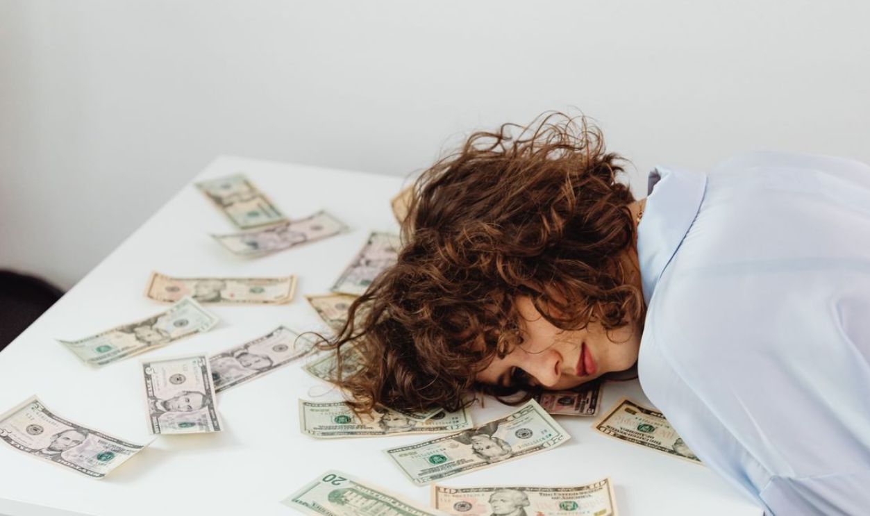 A woman with curly hair resting her head on a table covered in US dollar bills