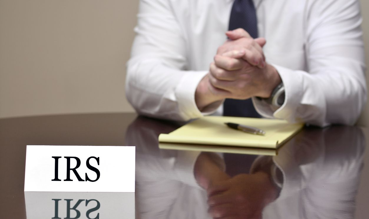 Businessman at desk with papers and card making hand gestures IRS Agent