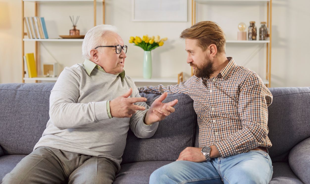 Young son and elderly father having conversation. White haired old man in his 60s or 70s sitting on sofa at home together with adult son, talking about life and sharing advice. Senior parents concept