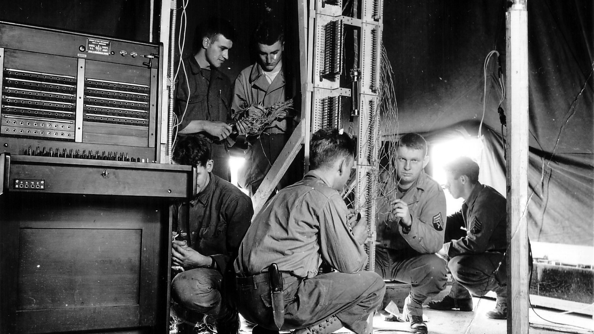 Sous une tente, des hommes de l'US Army Signal Corps installent un standard téléphonique Switchboard 58.
De face un T/4, derrière un autre T/4 près de la sortie de la tente.
De dos avec le couteau à la ceinture un T/3.
Reportage sur l'installation de deux