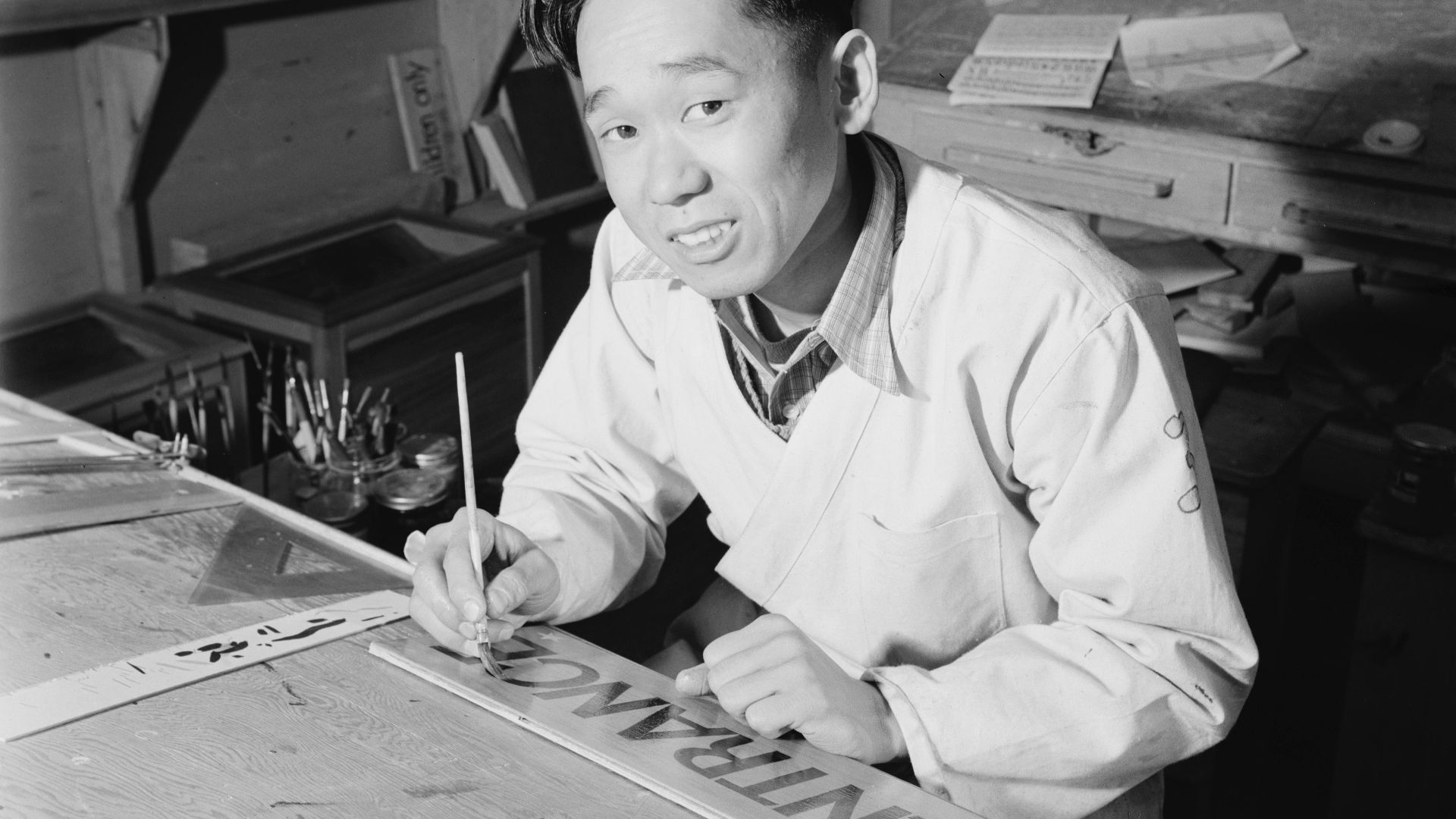 Original caption: Akio Matsumoto, commercial artist. Akio Matsumoto, half-length portrait, seated at desk, facing front, painting sign.