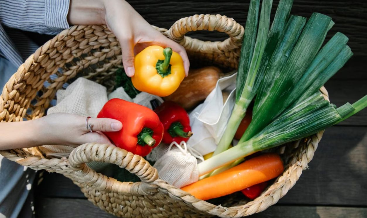 Woven basket with fresh vegetables including bell peppers and leeks held by hands outdoors