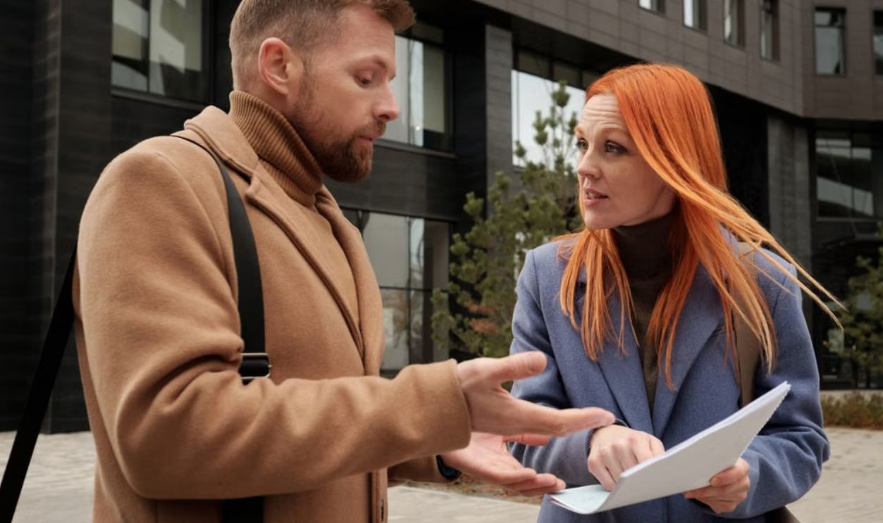 A man and woman in coats discussing documents outside a modern building