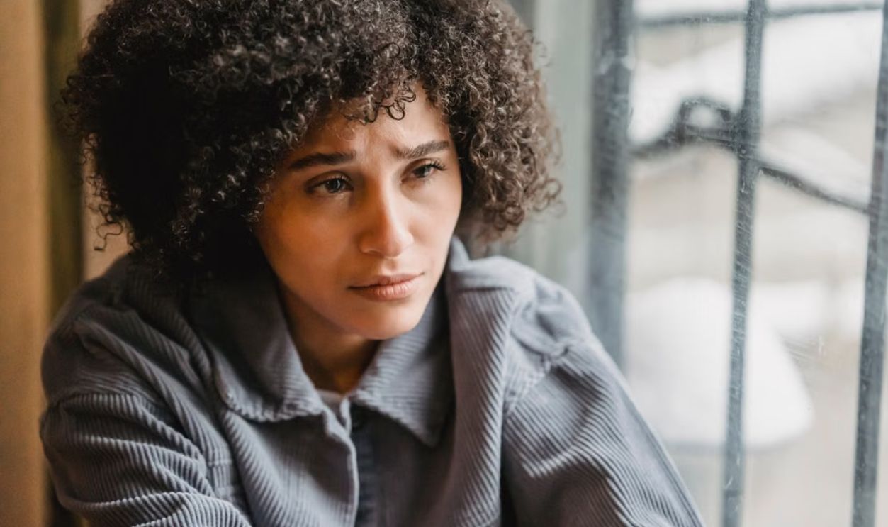 A young woman with curly hair sits pensively by a window, reflecting emotions indoors.