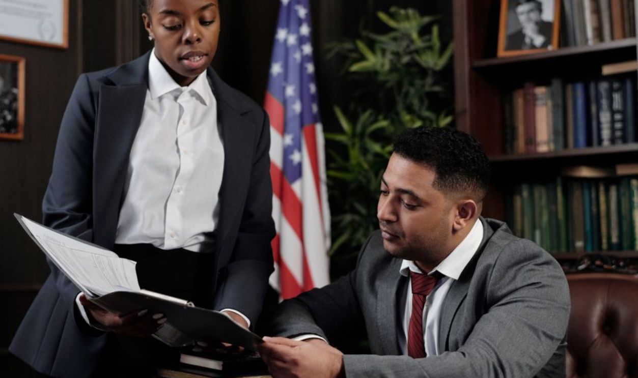 Two professionals discussing documents in an office setting with an American flag in the background