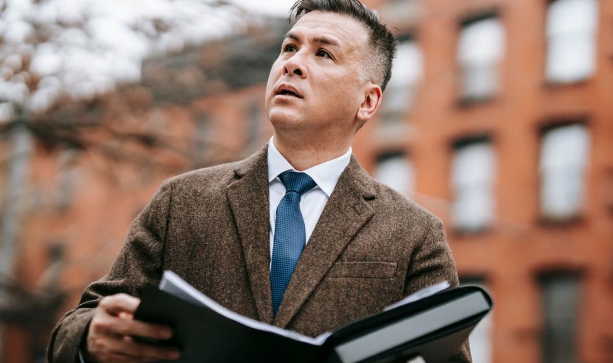 A professional man in a formal coat looks upwards while holding a folder outdoors