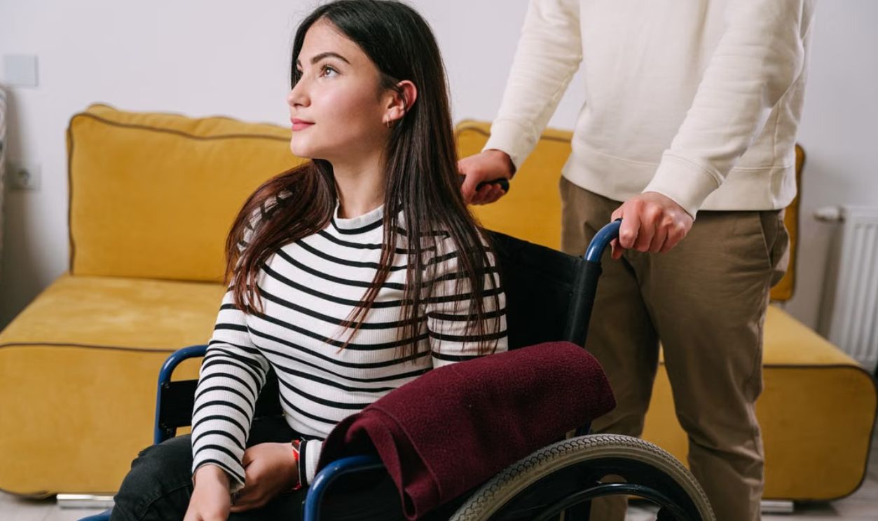 A young woman in a wheelchair being assisted indoors by a caregiver, showcasing support and mobility