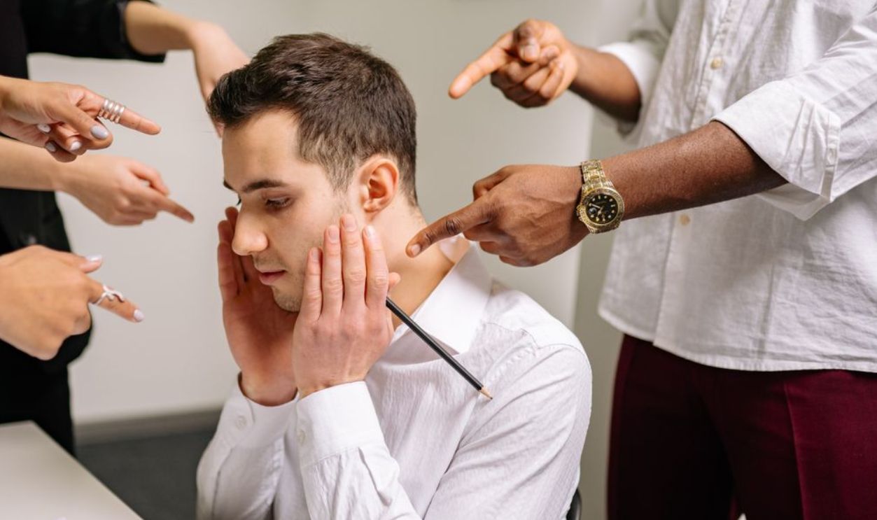 A stressed man at the office surrounded by pointing fingers, representing workplace bullying and stress