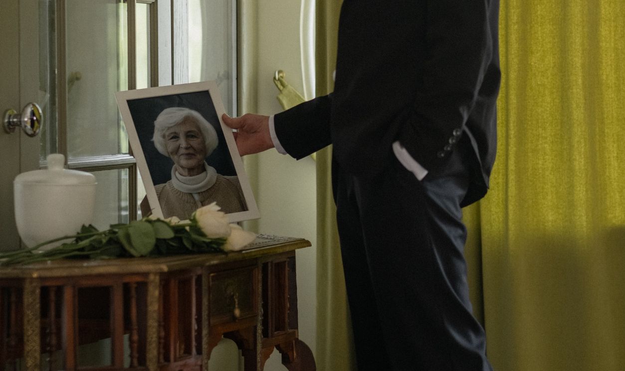 A man in mourning stands indoors reflecting on the photo of a loved one