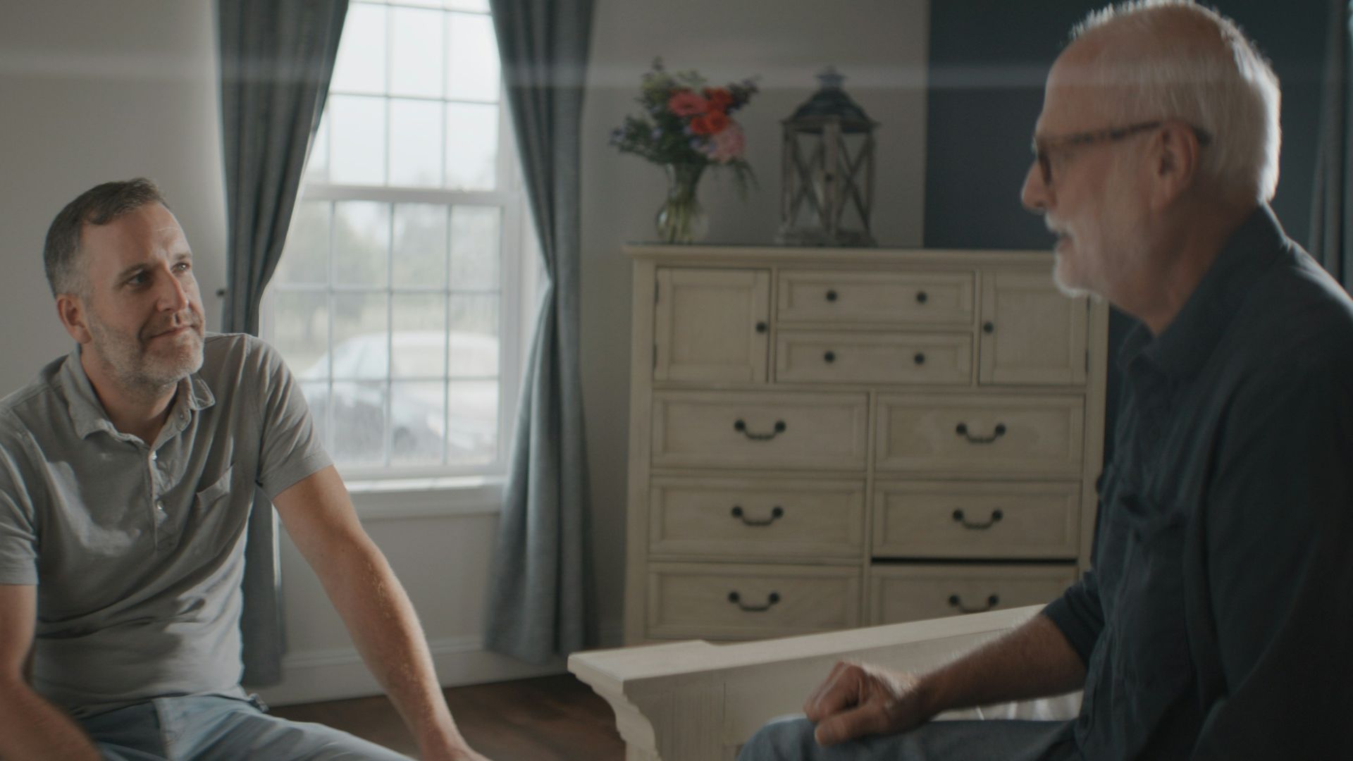 Two men conversing in a sunlit bedroom.