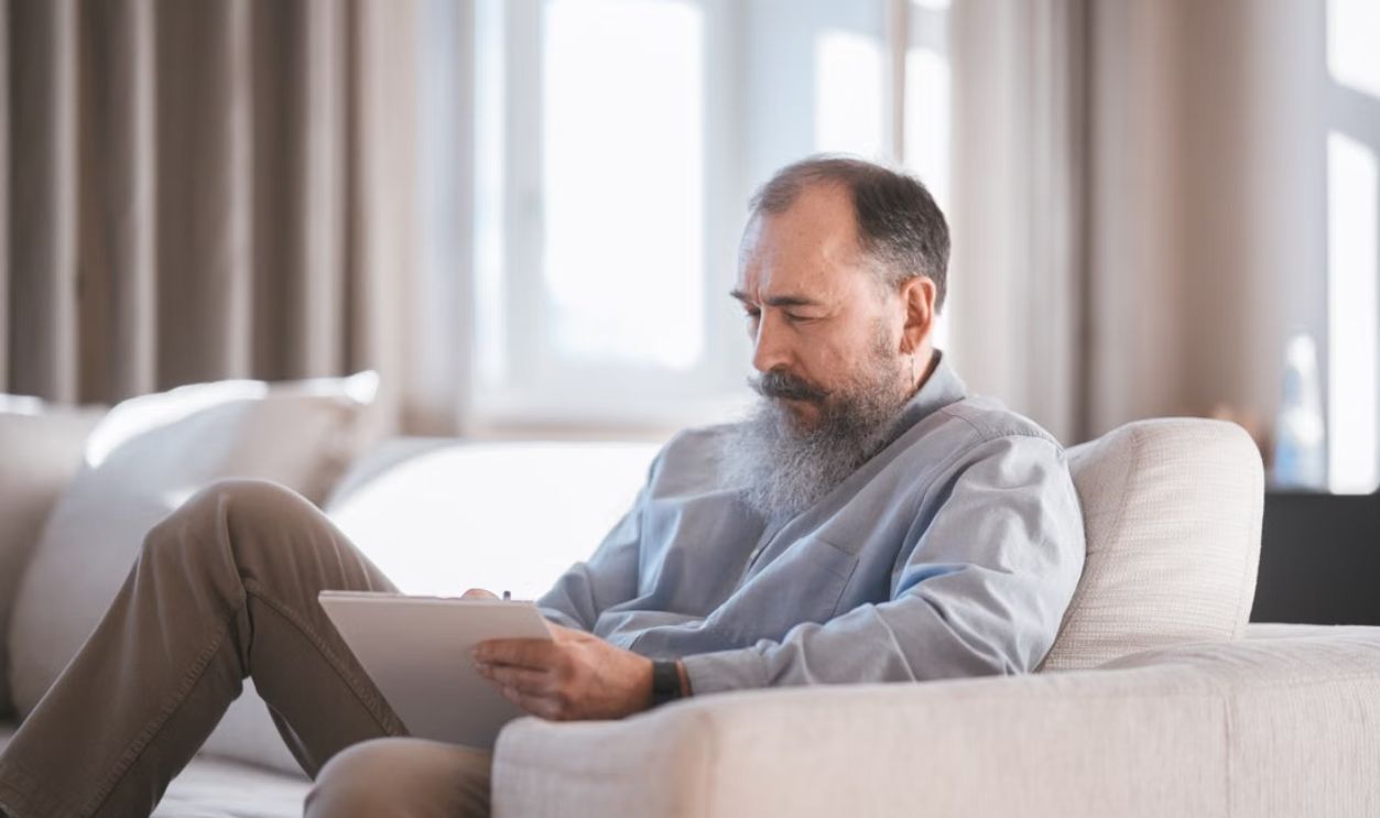 Elderly man sitting on sofa writing on a tablet with focus and concentration