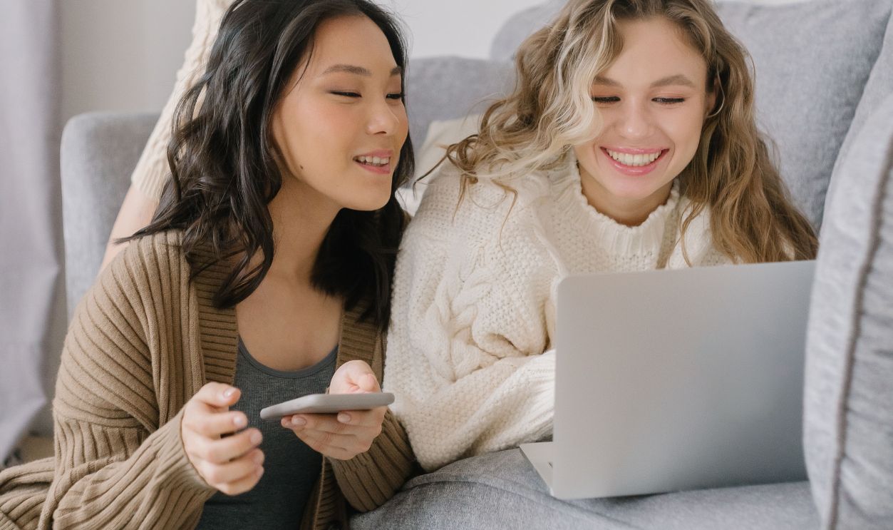 Two women laughing while using a laptop and smartphone in a cozy living room setting