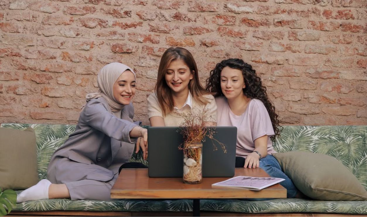 Three diverse women smiling, collaborating on a laptop in a cozy café with a rustic brick wall backdrop
