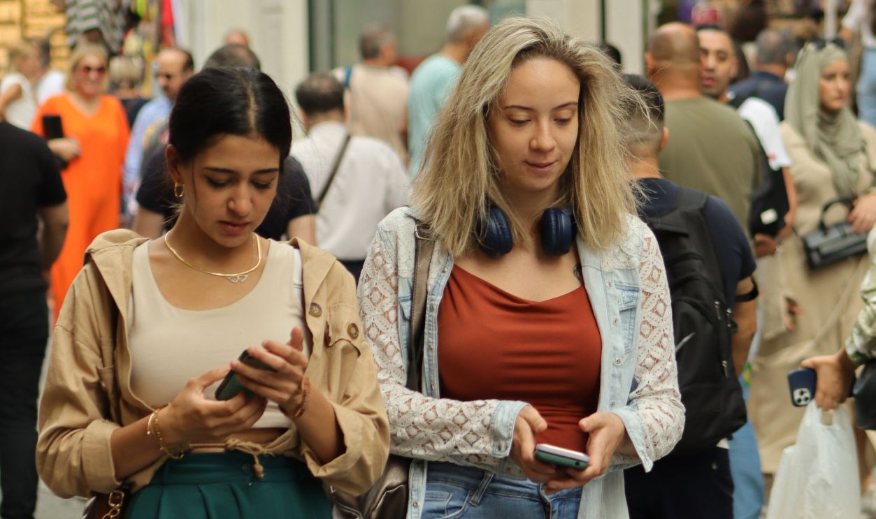 Two young women walking through a busy urban street, engaged with their smartphones.