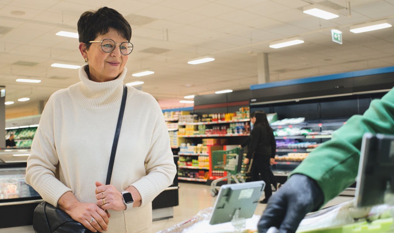 An older woman wearing a turtleneck sweater and glasses is buying fish at the supermarket fish counter, while the fishmonger selects the fish.